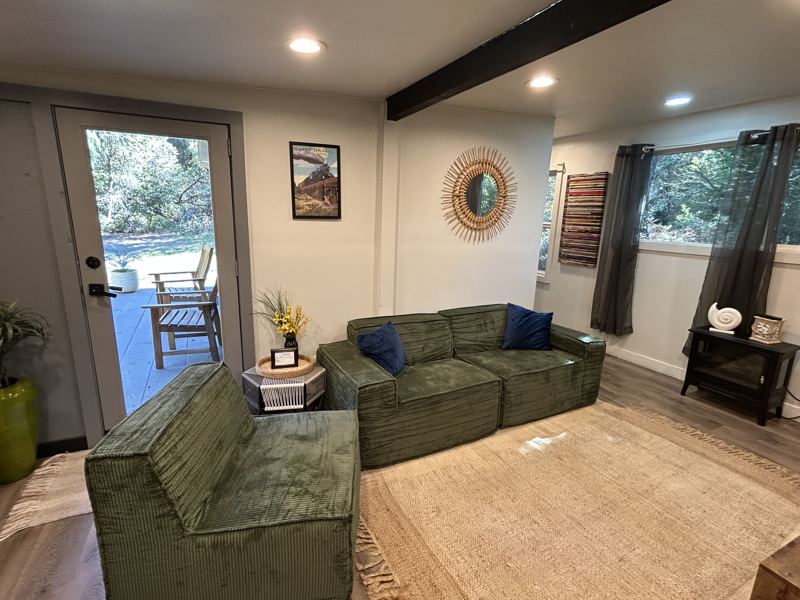 Living room with a green corduroy sofa and chair on a jute rug, a sunburst mirror and Skunk Train poster, and a glass door opening to the deck with Adirondack chairs among the trees